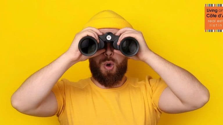 Man in yellow shirt and beanie looking through binoculars, symbolizing guided property search on the French Riviera.