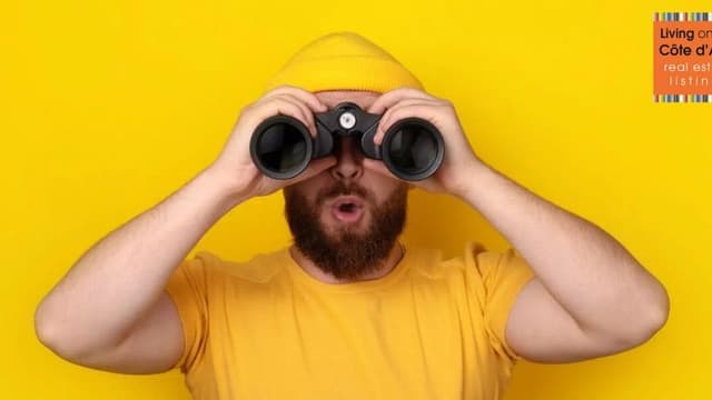 Man in yellow shirt and beanie looking through binoculars, symbolizing guided property search on the French Riviera.